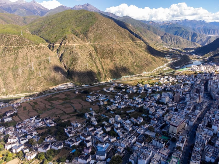 An aerial view of a mountainous village with white buildings, green fields, and a winding river, surrounded by mountains and a clear blue sky.