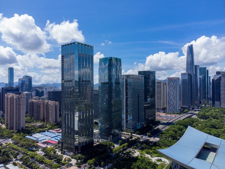 The image showcases a bustling cityscape with tall buildings, including skyscrapers and office buildings, against a backdrop of a clear blue sky with white clouds. The buildings vary in height and design, with some having reflective glass facades. The city is densely populated with roads and highways, and there are green trees scattered throughout. The image captures the essence of urban life, with the city's architecture and infrastructure.