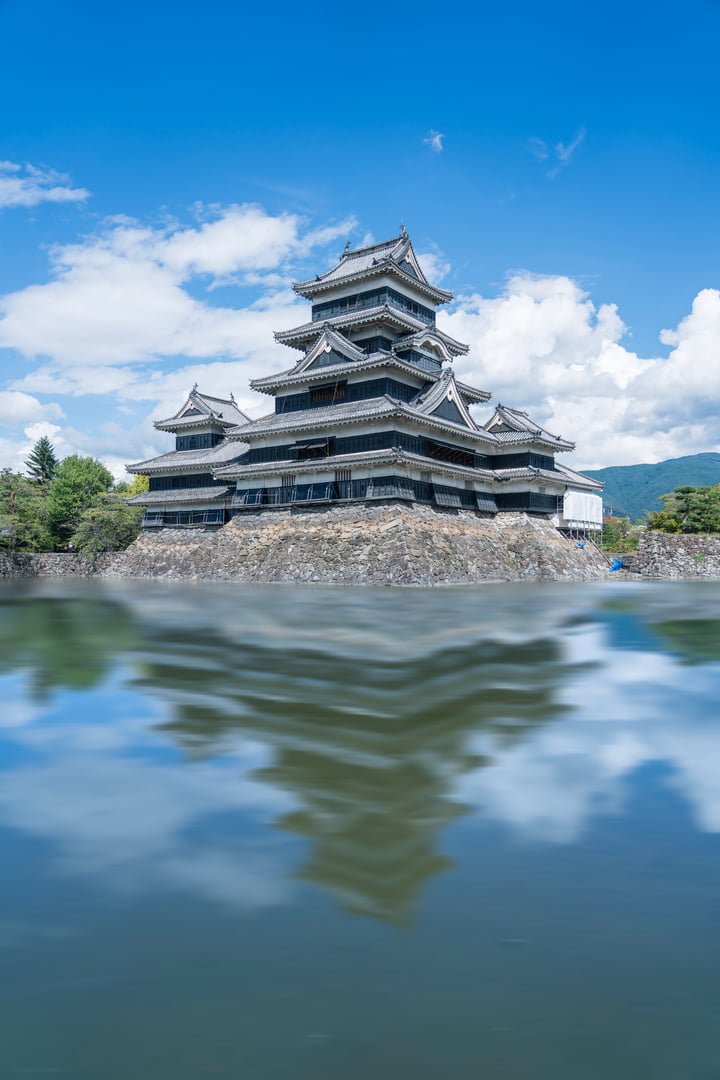 A majestic, multi-level castle-like structure with a gray and white color scheme is situated on a rocky outcropping, surrounded by a serene body of water. The castle's reflection is visible in the water, and the sky above is a clear blue with scattered clouds.