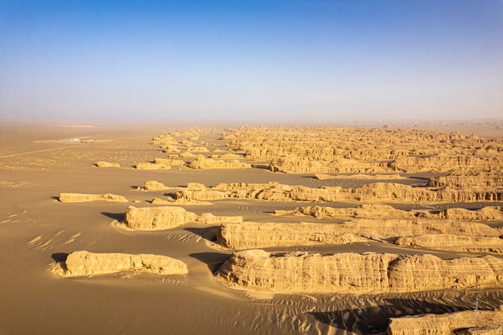 Aerial view of a desert landscape with sandy formations, a clear blue sky, and a few distant structures.