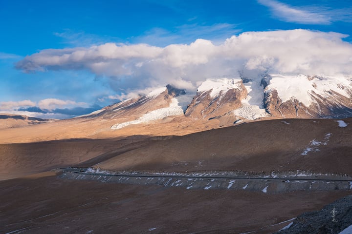 A breathtaking view of a mountain range with snow-capped peaks, set against a blue sky with white clouds, is captured in the image. The mountains are brown and tan, with patches of snow on their peaks. A winding road cuts through the landscape, leading the viewer's eye towards the mountains. The image is taken from a high vantage point, emphasizing the grandeur of the scene.