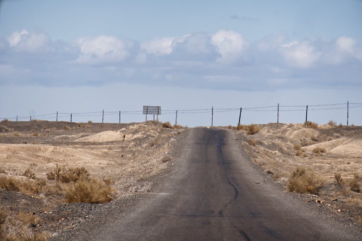 A solitary, winding black road stretches into the distance, bordered by barren, dry grass and a fence of barbed wire. The sky above is a canvas of blue, dotted with fluffy white clouds.