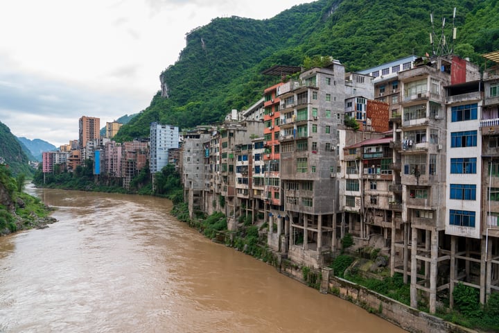 A cityscape with colorful buildings on a riverbank, surrounded by mountains and a cloudy sky.