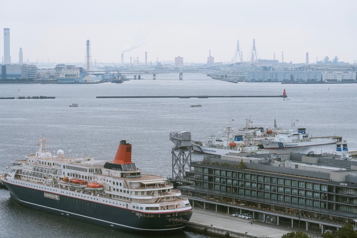 A large cruise ship with a red funnel is docked at a harbor, surrounded by a cityscape with tall buildings and cranes.