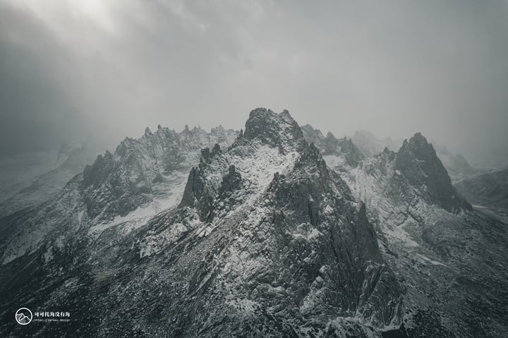 A black and white photograph captures a mountain range under a cloudy sky, with snow-covered peaks and a rocky mountain in the foreground.