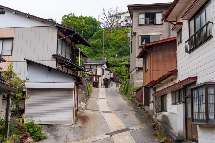 A narrow, steep street lined with white and brown houses, with a garage and a white house on the right side, surrounded by greenery and a hill in the background.