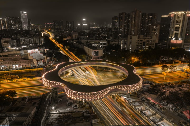 A large, circular sports complex is surrounded by a bustling cityscape, with cars and buildings in the background.