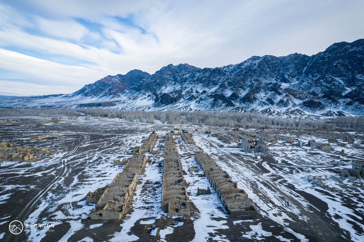 An aerial view of a snow-covered mountain range with a few buildings in the foreground, surrounded by a vast expanse of snow and a clear blue sky with white clouds.