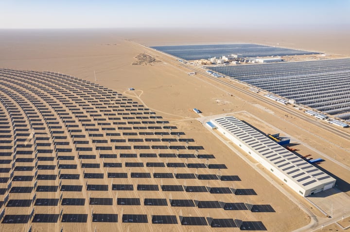 An aerial view of a large solar farm in a desert-like environment, with solar panels arranged in a circular pattern and a large white sun tower in the center. The farm is surrounded by a vast expanse of sandy terrain and a few wind turbines in the distance.