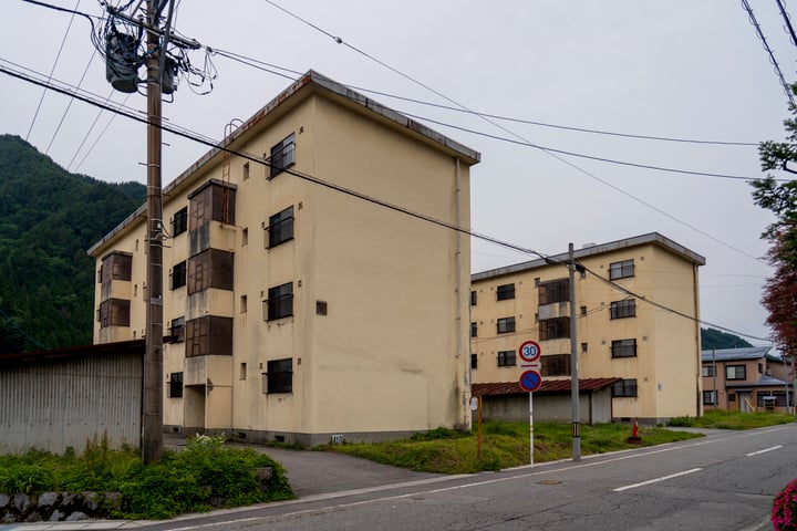A large tan building with brown windows and balconies, a red fire hydrant, and a blue and white traffic sign on a wooden pole are on a street corner.