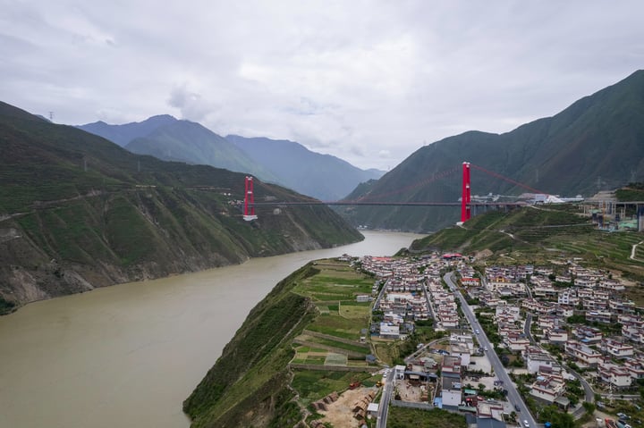 A red suspension bridge spans a river in a valley, with a city on the left bank and mountains in the background.