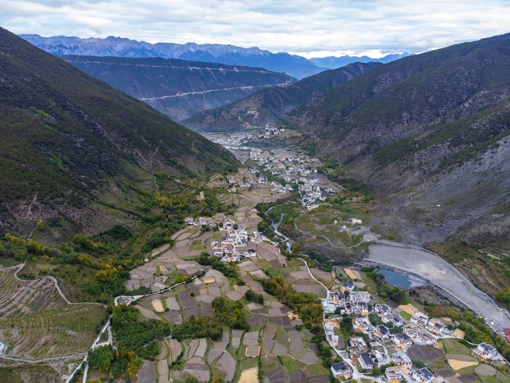 An aerial view of a valley with a winding river, surrounded by mountains, houses, and fields, with a clear blue sky above.