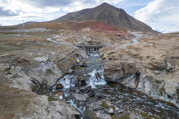 A serene landscape features a small stream flowing through rocky terrain, with a bridge connecting two sides. A mountain with patches of red and brown soil serves as a backdrop.