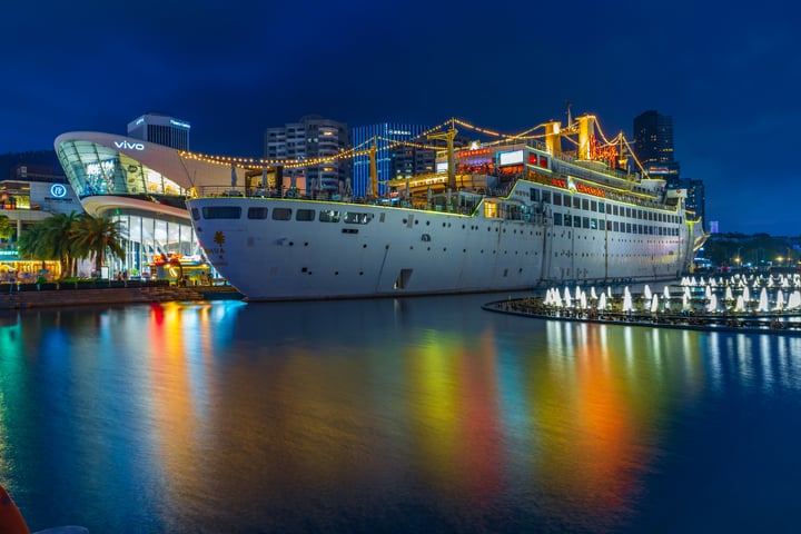 A large white cruise ship with a yellow crane on its side is docked in a harbor, surrounded by a calm body of water. The city skyline with tall buildings and a crane is visible in the background.