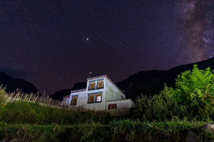 A white two-story house with blue accents is nestled in a verdant landscape, surrounded by lush greenery and mountains. The house is illuminated by a starlit night sky, with the Milky Way visible in the distance.