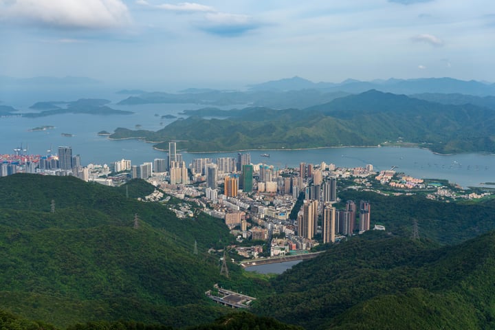 A bustling city nestled amidst verdant mountains and a large body of water, with tall buildings and a bridge spanning the water.