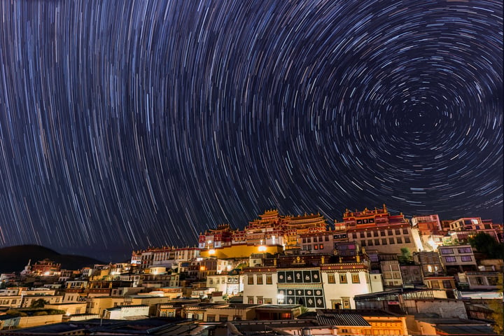 A starry night scene in a mountainous city features a circular pattern of white stars streaking across the sky, creating a mesmerizing effect. The cityscape is densely packed with buildings, some with orange roofs, and a few with red roofs. The buildings are illuminated, casting a warm glow against the night sky. The image is taken from a high vantage point, offering a panoramic view of the city and its surroundings.