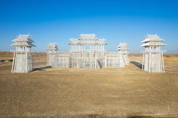 An aerial view of a white, pagoda-like structure with multiple levels and a central roof, surrounded by a barren desert landscape.