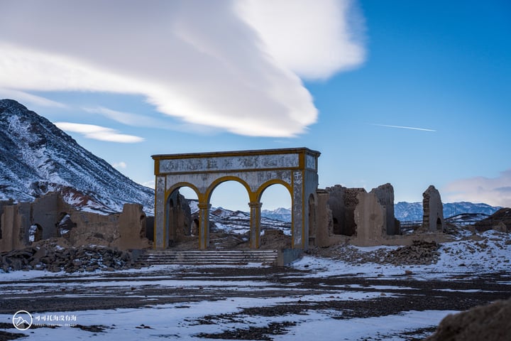 An old, weathered stone archway stands in a barren landscape, surrounded by snow and mountains, with a clear blue sky above.