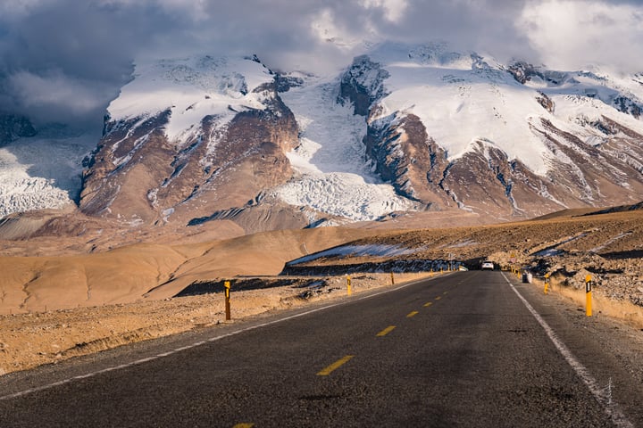 A black road with yellow lines cuts through a barren landscape, with snow-capped mountains in the background. A solitary car drives along the road, and a yellow post stands on the right side. The sky is filled with clouds, casting a soft light on the scene.