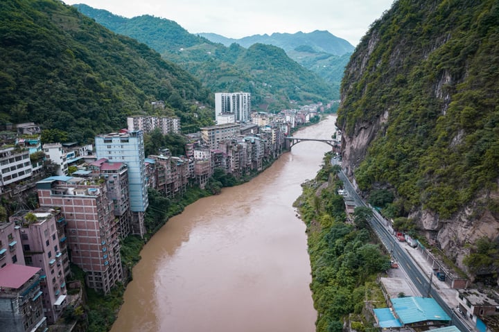 A serene mountain landscape features a winding river, a bridge, and a cluster of buildings on the left side, with a mountain range in the background.