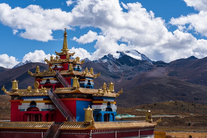 A mountain range with snow-capped peaks and a blue sky with white clouds is the backdrop of a colorful, multi-level pagoda-like structure. The structure is surrounded by a grassy area and a fence, with a small pond in the foreground.