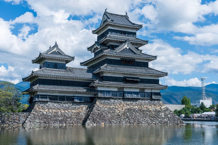 A majestic, multi-level Japanese castle stands on a stone wall, surrounded by a serene body of water. The castle's gray and white facade contrasts with the blue sky, while the mountains in the distance add depth. The image captures the castle's grandeur and tranquility, with no discernible text or actions.