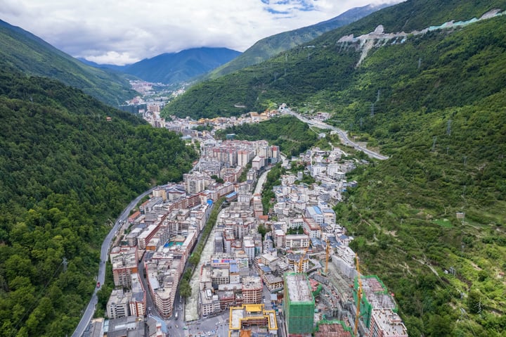 An aerial view of a mountain town nestled between verdant green mountains, with a winding river running through the center, surrounded by towering green mountains. The town is densely populated with buildings of varying heights and colors, and construction cranes are visible on the right side. The sky is partly cloudy, casting a soft light over the scene.
