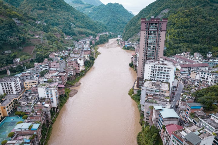 An aerial view of a river with a brownish riverbed, surrounded by lush green mountains and a tall, red-brick building.