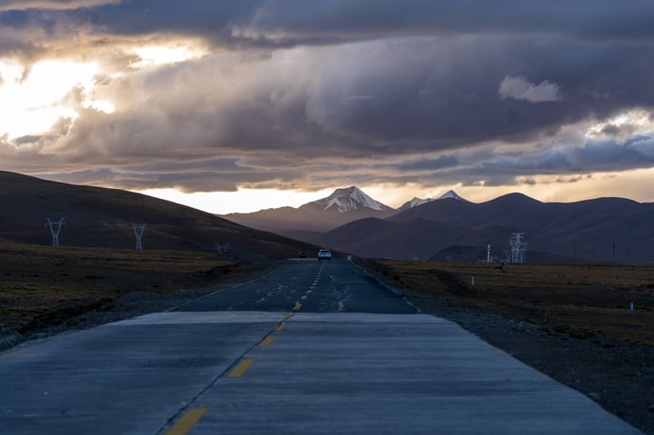 A long, empty road stretches into the distance, with a mountain range in the background and a cloudy sky overhead.