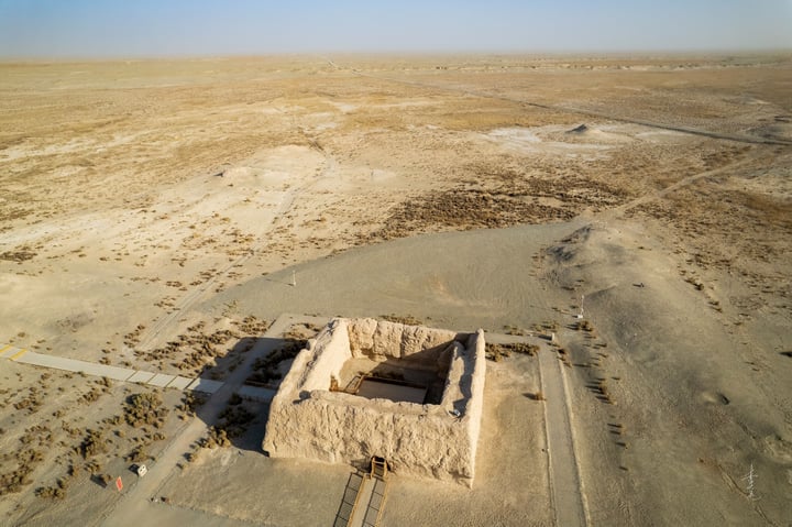 An aerial view of a desert landscape with a small, rectangular stone structure in the center, surrounded by a vast expanse of dry, sandy terrain. The sky is clear blue, and the ground is covered in dirt and rocks.