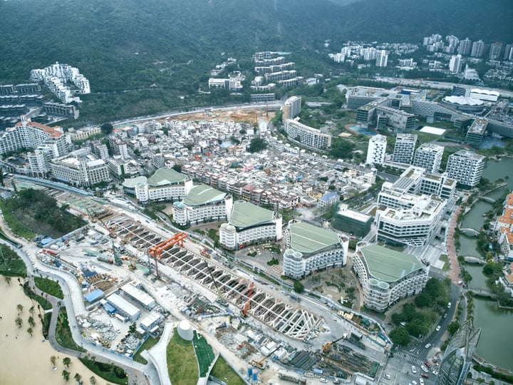 An aerial view of a city with a river, surrounded by mountains, shows a mix of modern and older buildings, with a large factory on the riverbank.