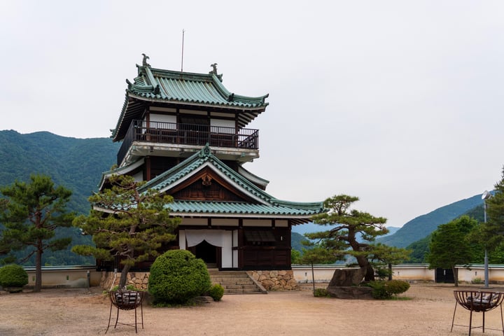 A traditional Japanese temple with a green roof and white walls stands in a serene landscape, surrounded by trees and shrubs. The temple is set against a backdrop of mountains and a cloudy sky, creating a harmonious blend of architecture and nature.