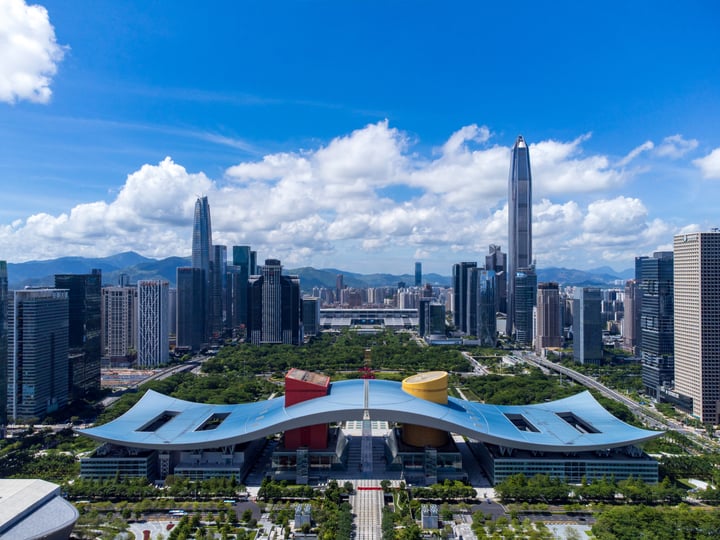 The image showcases a bustling cityscape with skyscrapers, a large, colorful building, and a park-like structure. The sky is a clear blue with white clouds, and the city is surrounded by mountains. The buildings vary in height and design, with some having curved roofs and others having straight roofs. The park-like structure is surrounded by trees and has a large, curved roof. The image captures the essence of urban life with a blend of architecture, nature, and cityscape.