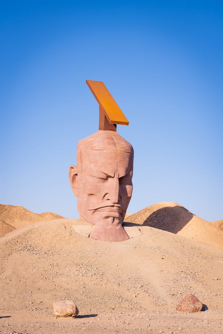 A large, red clay sculpture of a man's head with a pointed nose and a raised eyebrow is situated on a sandy hill in a desert-like environment. The sculpture is surrounded by small rocks and is positioned in the center of the image.