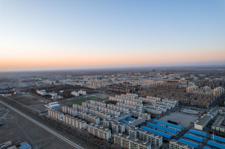 An aerial view of a densely populated city with a mix of modern and older buildings, surrounded by a vast expanse of open space and a road. The sky is a mix of blue and orange hues, indicating either sunrise or sunset.