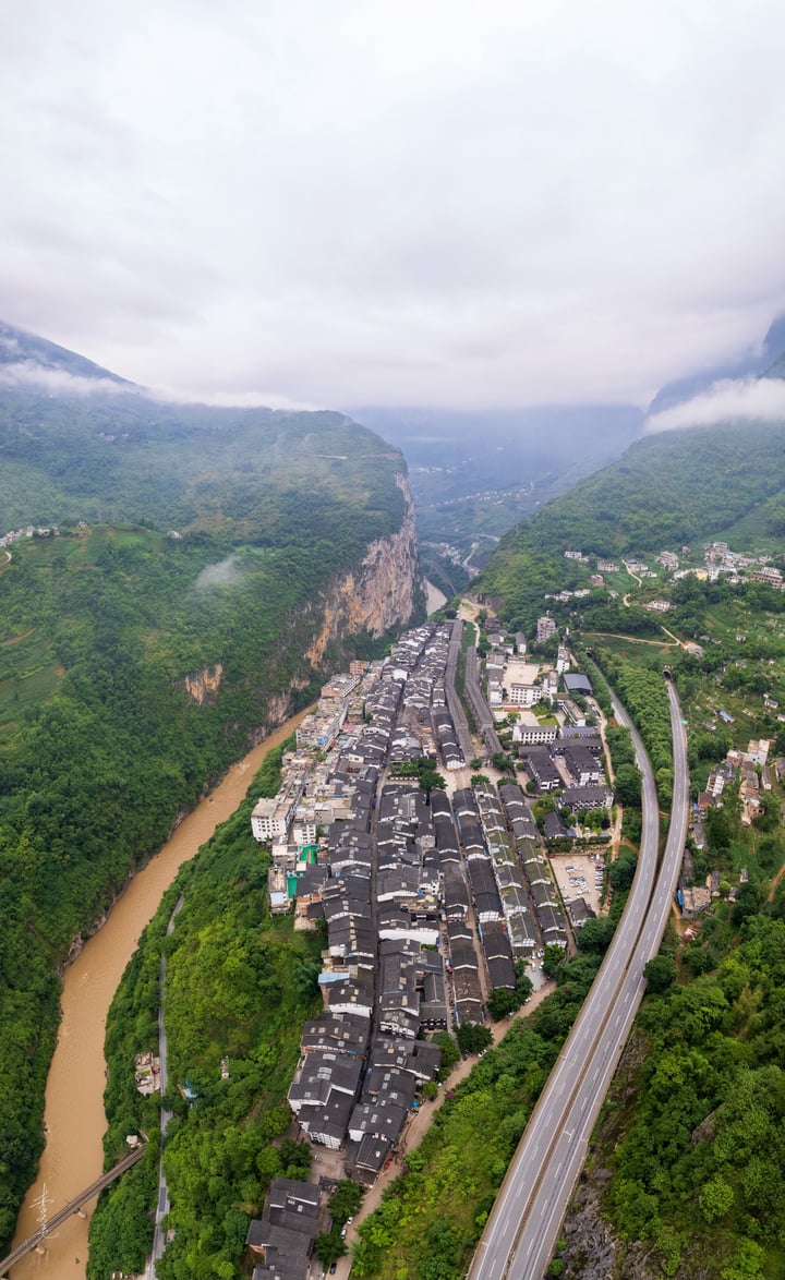 An aerial view of a mountain town nestled between verdant hills and a winding river, with a road running through the center, surrounded by lush greenery.