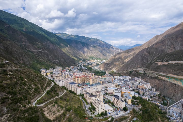 A serene valley city with white buildings, blue roofs, and green mountains, surrounded by a river and a winding road, is captured from a high vantage point.