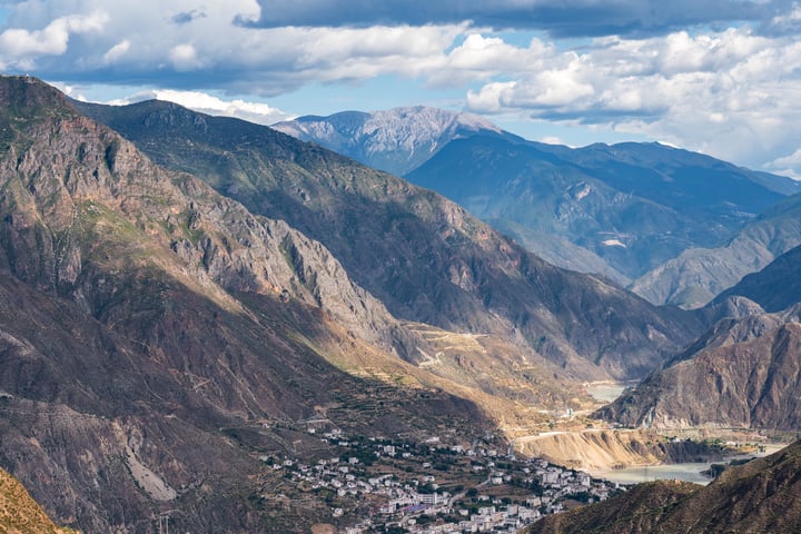 A breathtaking mountainous landscape with a small town nestled amidst towering mountains, surrounded by a river and a clear blue sky with white clouds.