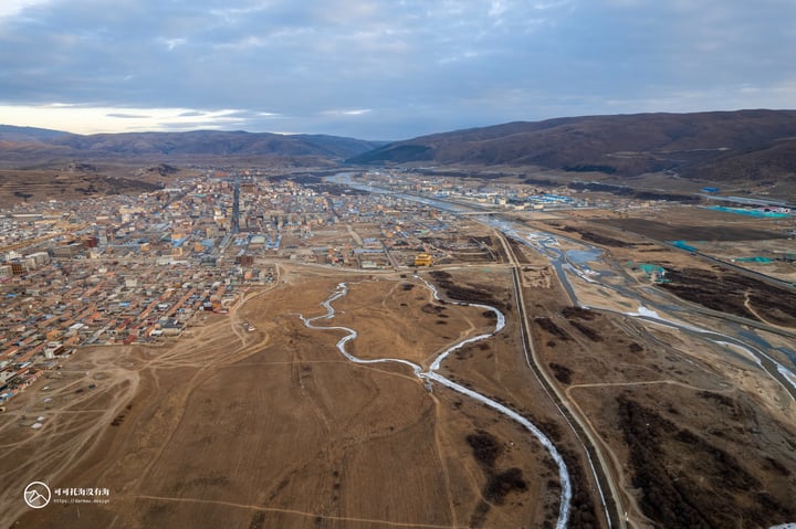 An aerial view of a city with a mix of buildings and mountains, surrounded by a river and a mountain range.