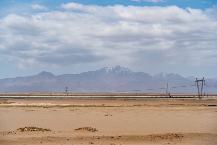 A vast desert landscape with a mountain range in the background, a power line in the foreground, and a clear blue sky with clouds.