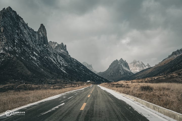 A long, empty road stretches into the distance, bordered by a mountain range with snow-capped peaks. The sky is filled with clouds, casting a soft light over the scene.