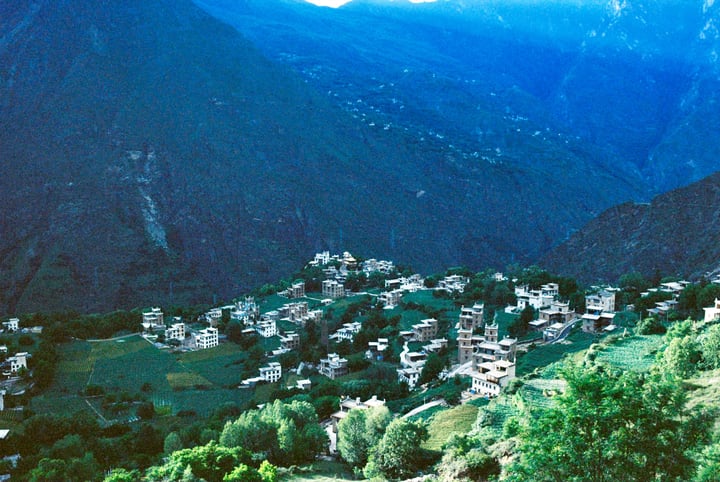 A picturesque mountain village nestled amidst verdant greenery, with white houses and trees, surrounded by snow-capped mountains.