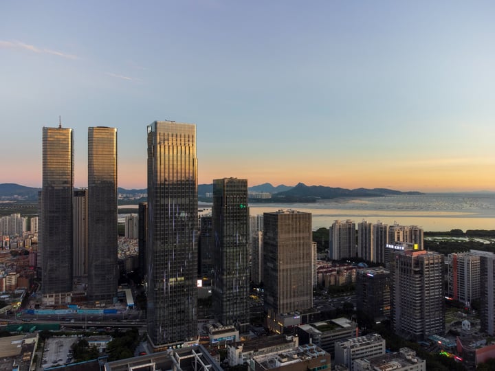 A cityscape with tall buildings, including skyscrapers, is captured during sunset, with the sun setting behind mountains and creating a warm, golden glow.