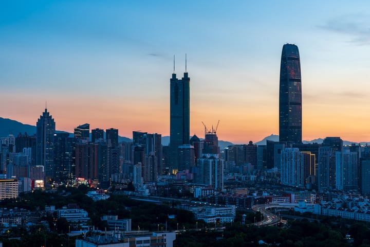 A city skyline at dusk, with a prominent skyscraper, a curved building, and a mountain range in the background.