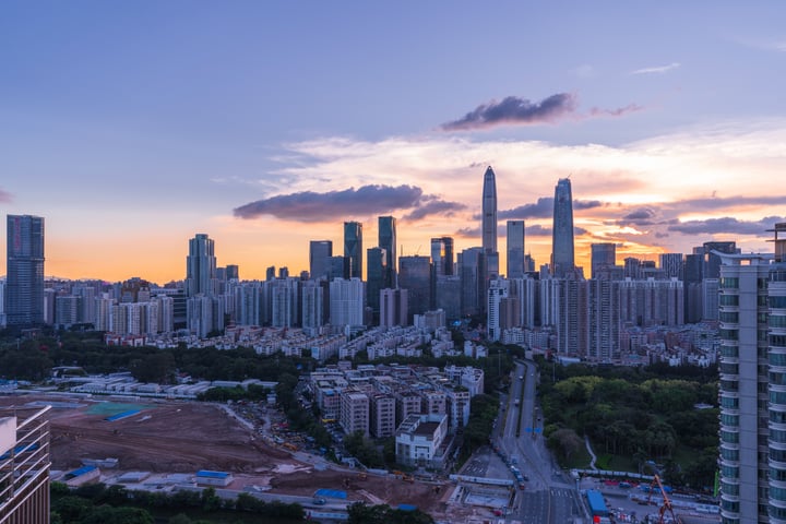 A city skyline at sunset, with tall buildings in various shades of gray, white, and blue, reflecting the warm hues of the setting sun. The city is bustling with activity, with a construction site visible in the foreground. The sky is a gradient of orange and blue, with clouds scattered across it.