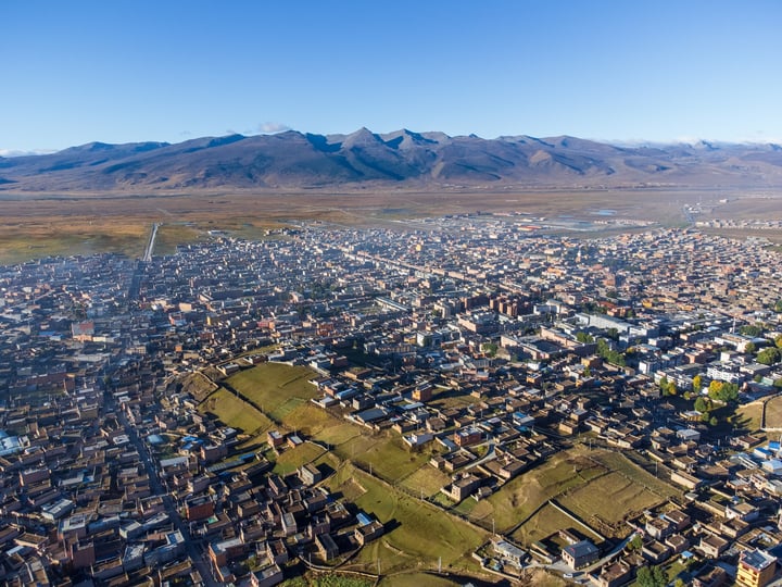 An aerial view of a city nestled between mountains, with a mix of buildings and green spaces, surrounded by snow-capped mountains.