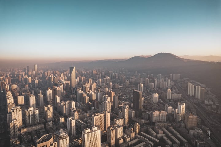 An aerial view of a bustling city with tall buildings, a mountain range in the background, and a warm, golden hue from the setting sun.