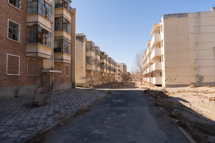 An empty, dusty alleyway with old brick buildings on both sides, surrounded by bare trees and a clear blue sky.
