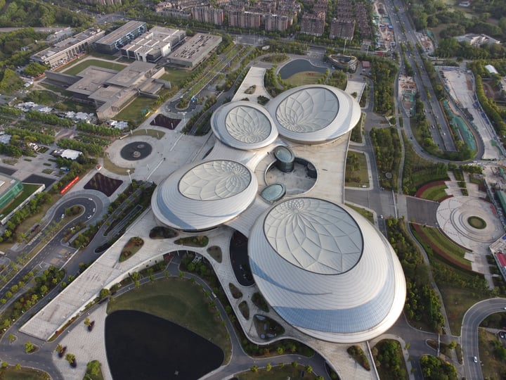 An aerial view of a modern, large, circular building surrounded by a park and a road, with a large circular pool in the center.